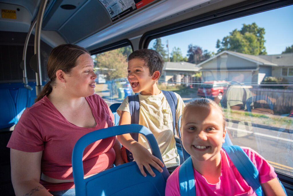 A woman and two children with backpacks sit together and smile on a city bus during daytime, with houses visible through the window.