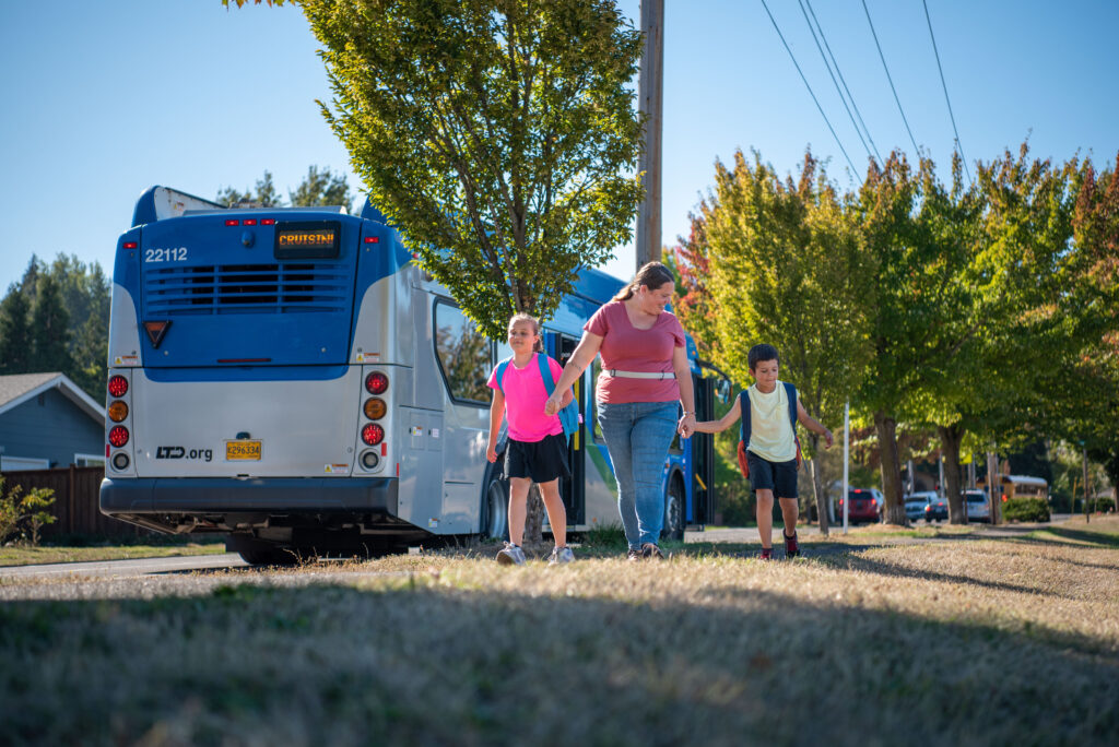 An adult and two children walk on a sidewalk near a stopped city bus on a suburban street lined with trees.