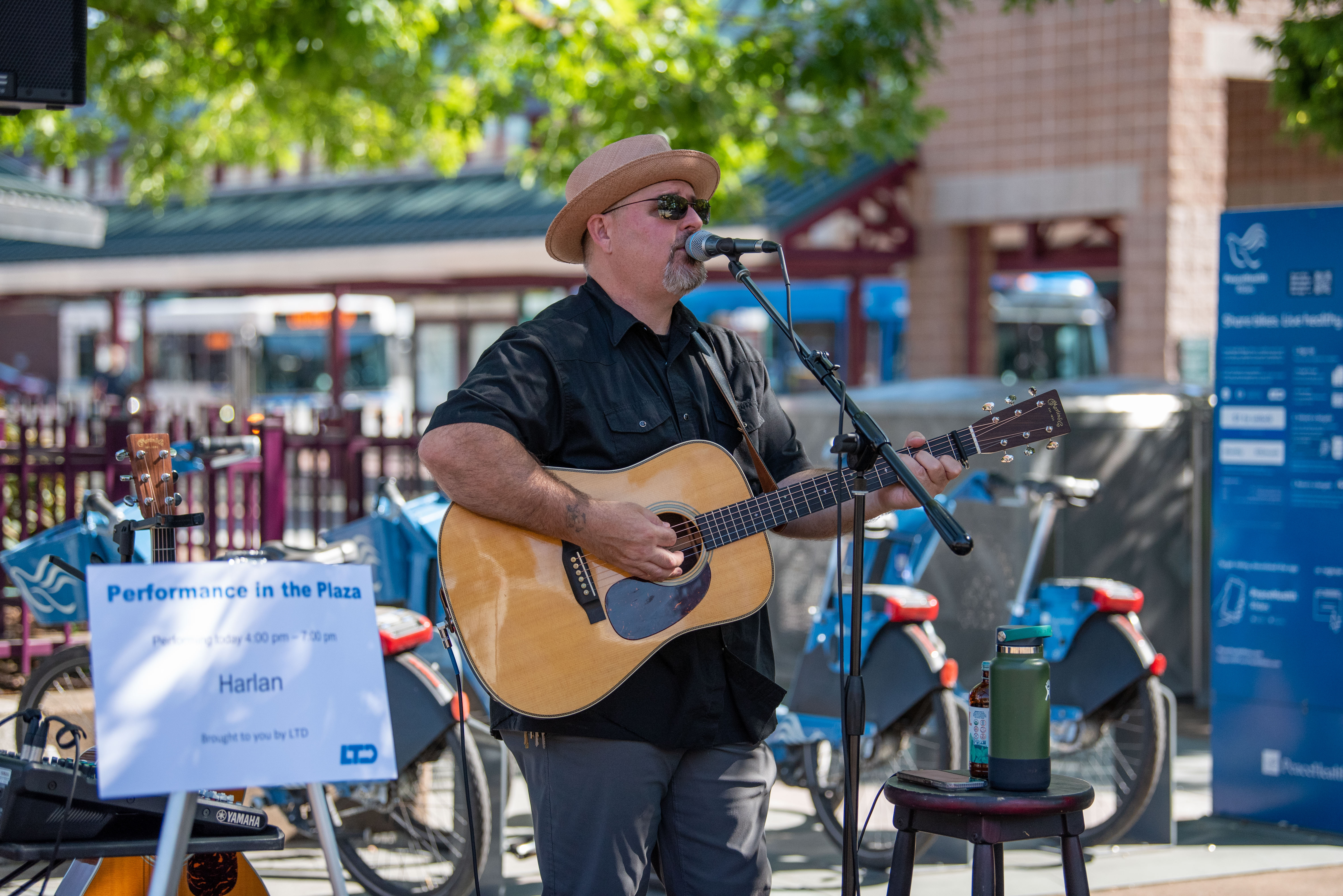 A musician wearing sunglasses and a hat plays an acoustic guitar and sings into a microphone at an outdoor event. A sign nearby reads "Performance in the Plaza" with "Harlan" listed.