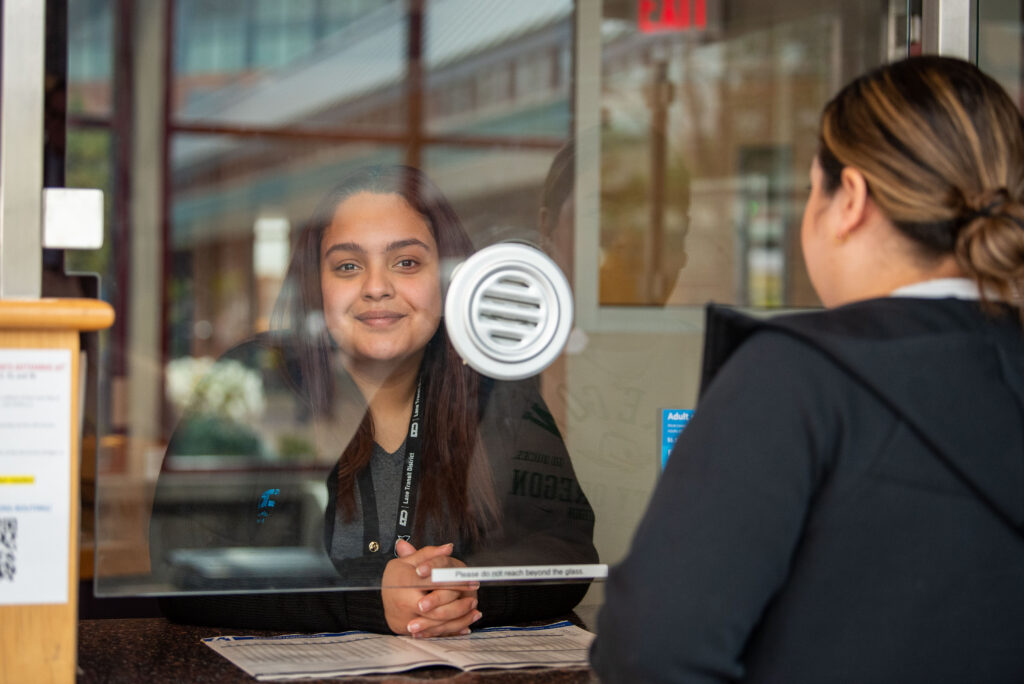 A woman sits behind a glass window at a service counter, facing another person on the other side, with papers and a sign visible on the counter.