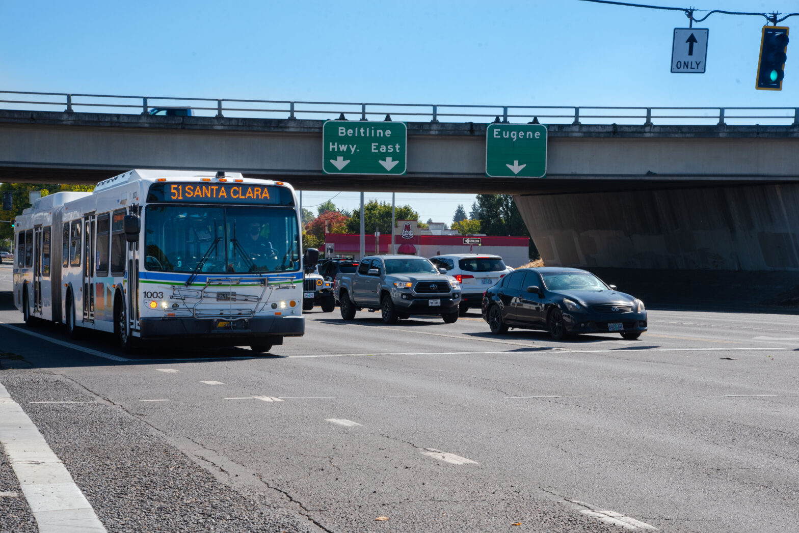 A city bus marked "51 Santa Clara" and several cars wait at a traffic light near a highway overpass with signs for Beltline Hwy. East and Eugene.