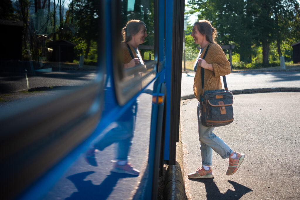 A woman with a shoulder bag stands next to a blue bus, her reflection visible in the bus window, on a sunny day.