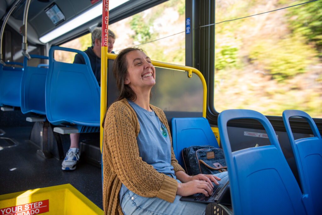 A woman sits on a bus with a laptop on her lap, smiling with her eyes closed. Blue seats and bus windows with blurred greenery are visible in the background.
