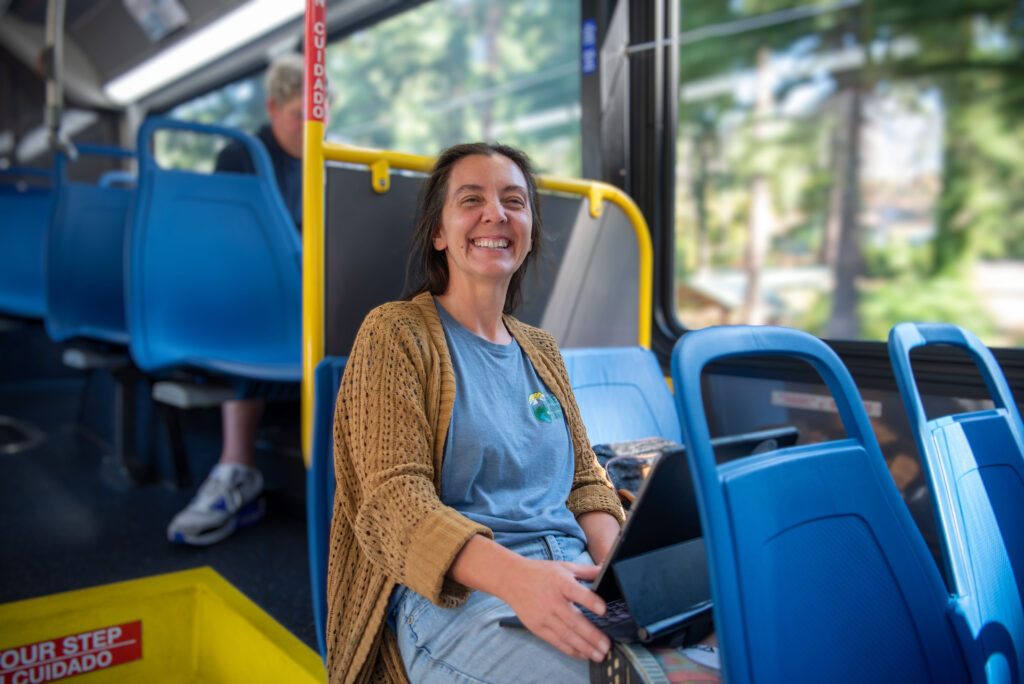 A woman sitting on a blue seat in a bus, smiling at the camera, with a laptop on her lap; trees and a blurred person are visible in the background.