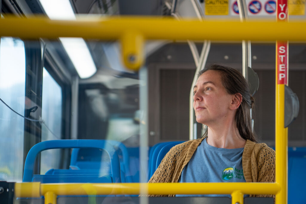 A woman with a blue shirt and brown cardigan sits alone on a bus, looking out the window. Blue seats and yellow rails are visible in the foreground.
