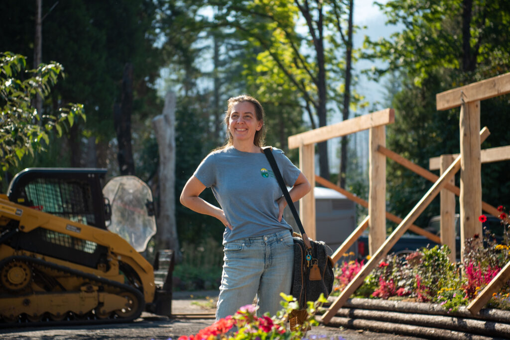 A woman in a gray T-shirt and jeans stands outdoors near a construction site, smiling, with a bag over her shoulder and flowers and equipment in the background.