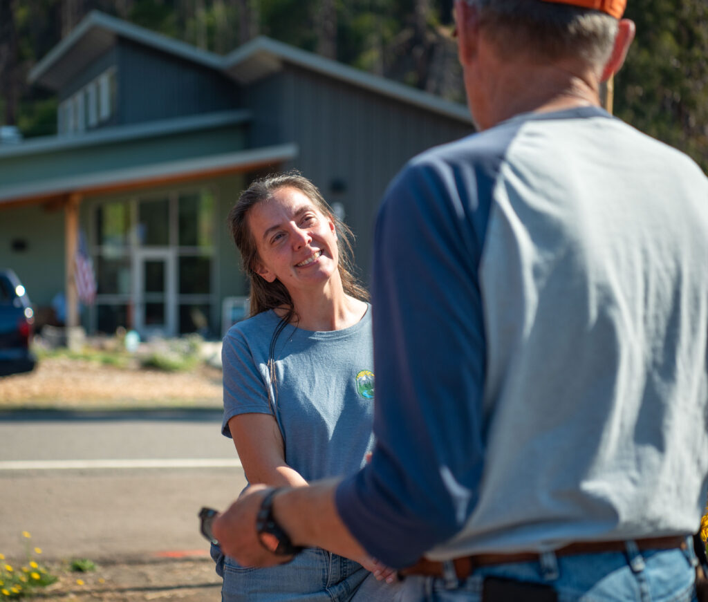 A woman in a blue shirt smiles and talks to a man outdoors, with a building and an American flag in the background.