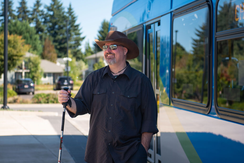 A man wearing sunglasses and a cowboy hat stands smiling beside a blue bus, holding a white cane. Trees and houses are visible in the background.