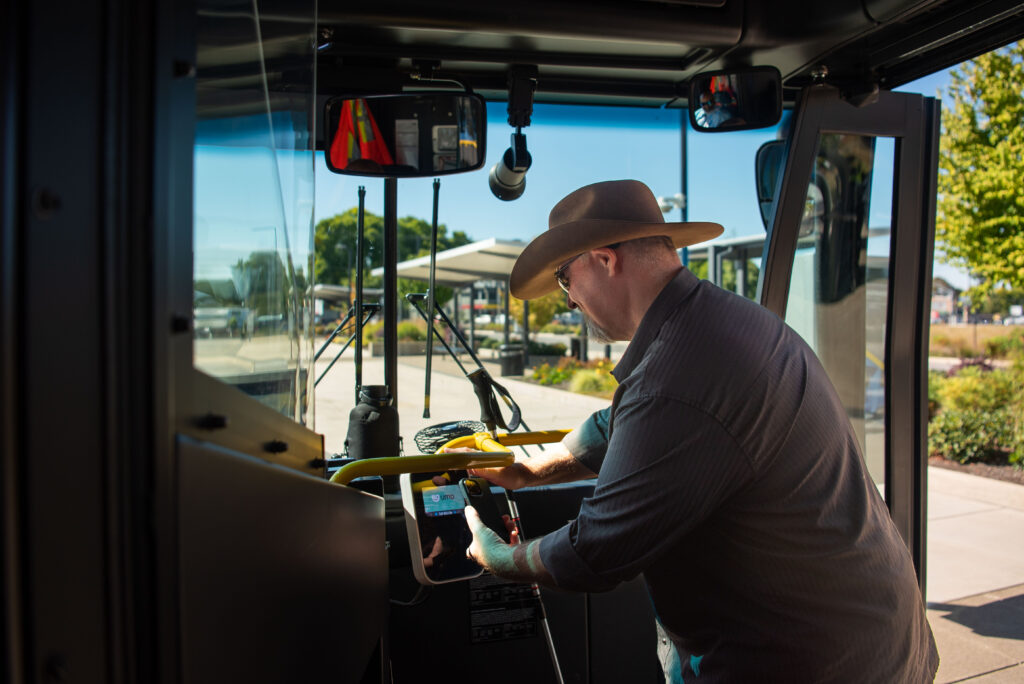A man in a cowboy hat stands inside a bus, using a device near the driver's controls on a sunny day.