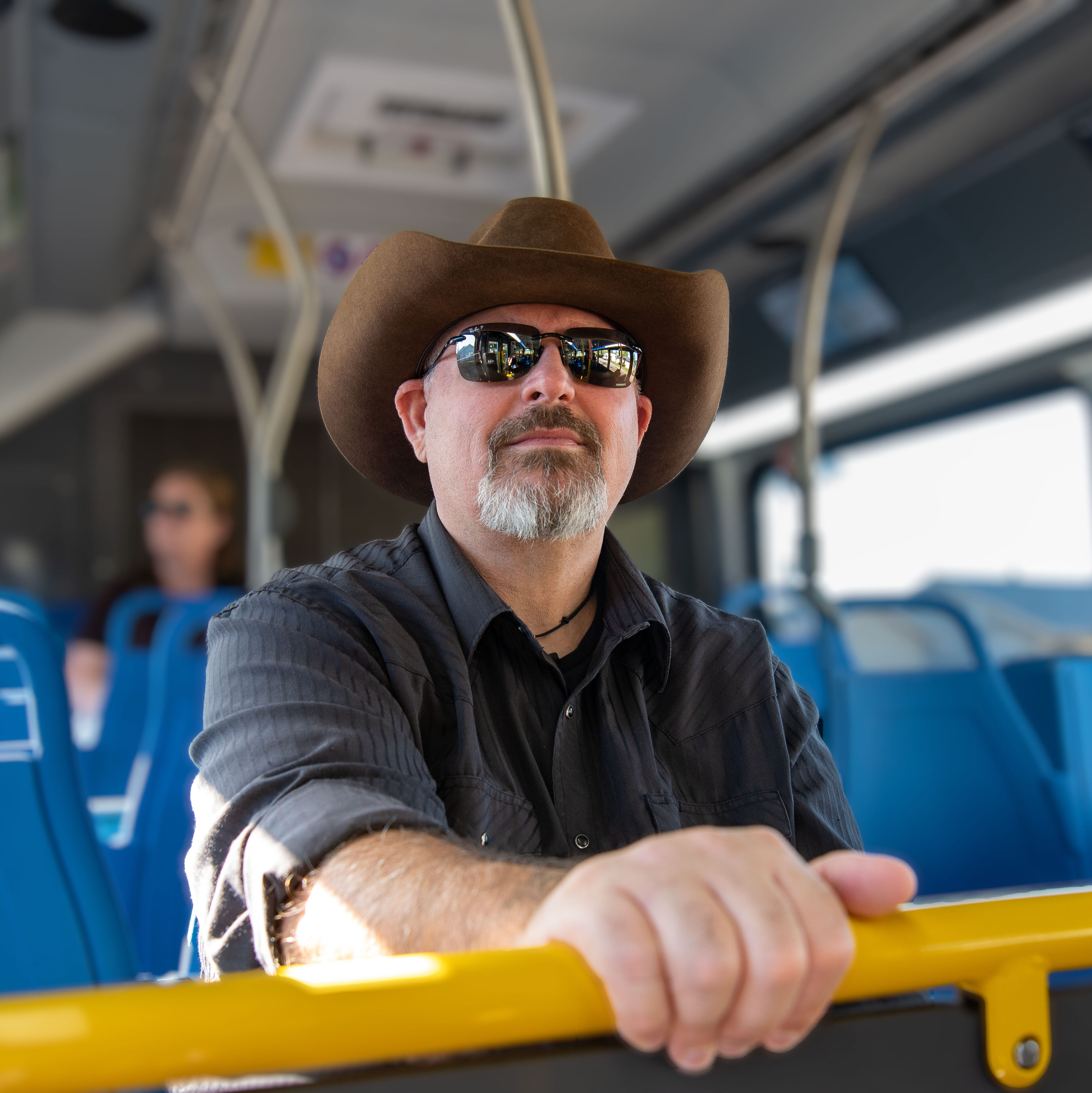 A man wearing a cowboy hat and sunglasses sits on a blue bus seat, holding onto a yellow railing, with other passengers visible in the background.