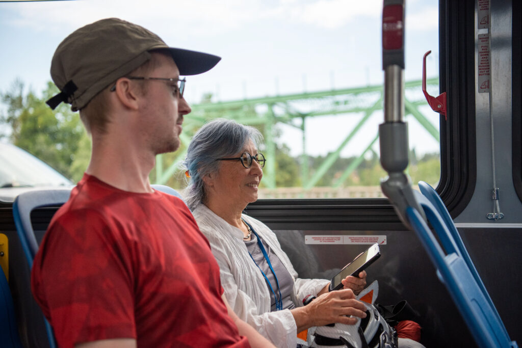 Two people sit side by side on a bus. The person in front wears a red shirt and cap, and the person behind, with gray hair, is smiling and holding a smartphone.