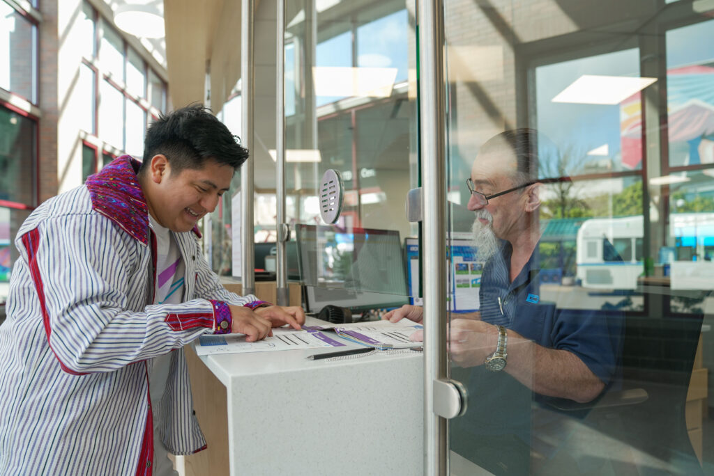 A man fills out paperwork at a counter while another man, seated behind a glass partition, assists him inside a modern office with large windows.