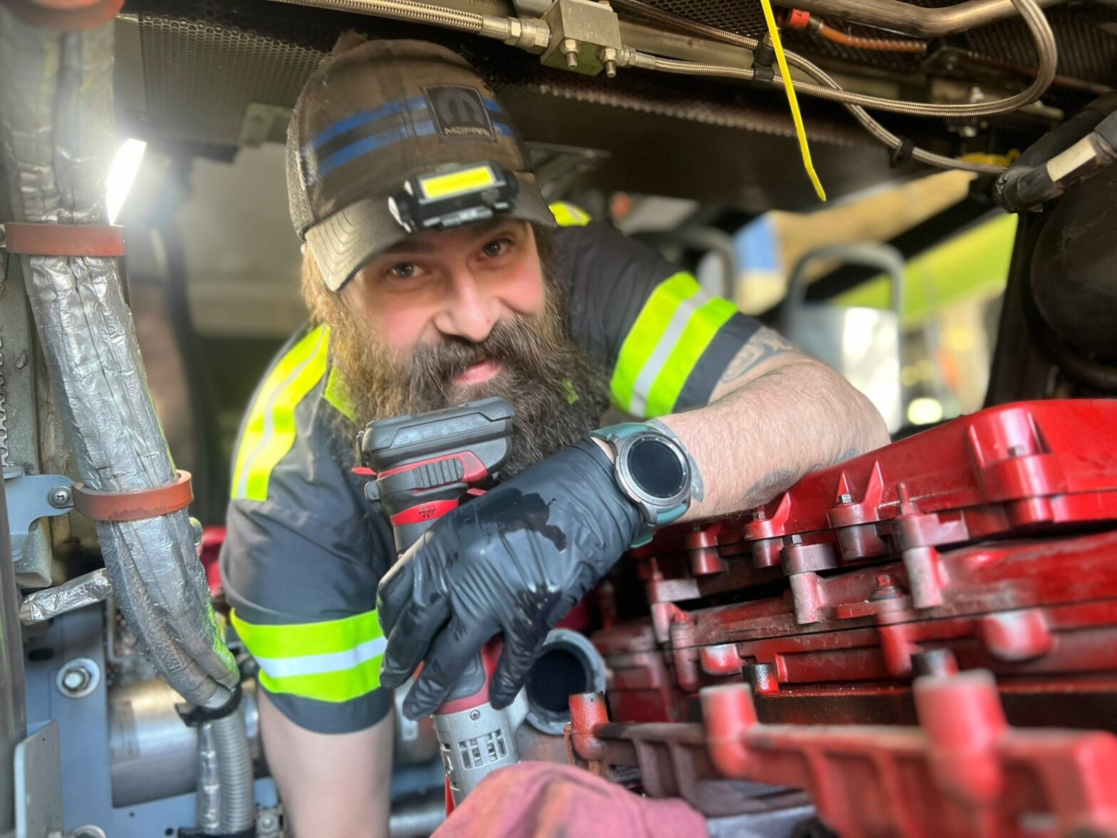 A mechanic wearing gloves and a cap works on machinery, holding a power tool and leaning over a red engine component.