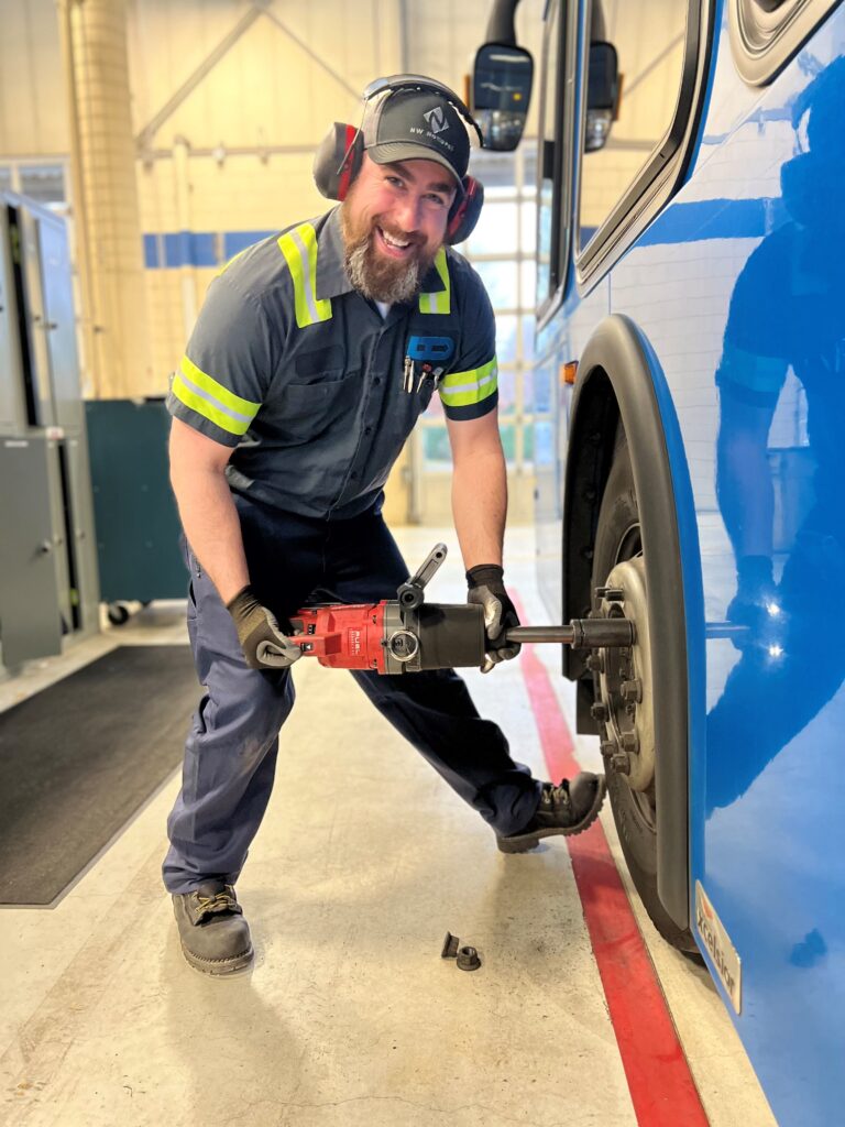 A mechanic wearing safety gear uses a power tool to work on the wheel of a blue bus inside a garage.