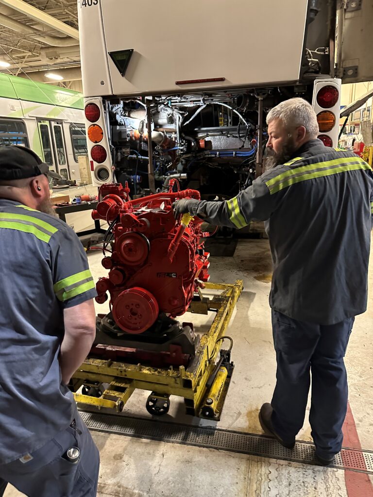 Two mechanics in uniforms stand by a red engine on a lift, preparing to install it into the rear of a bus inside a maintenance garage.