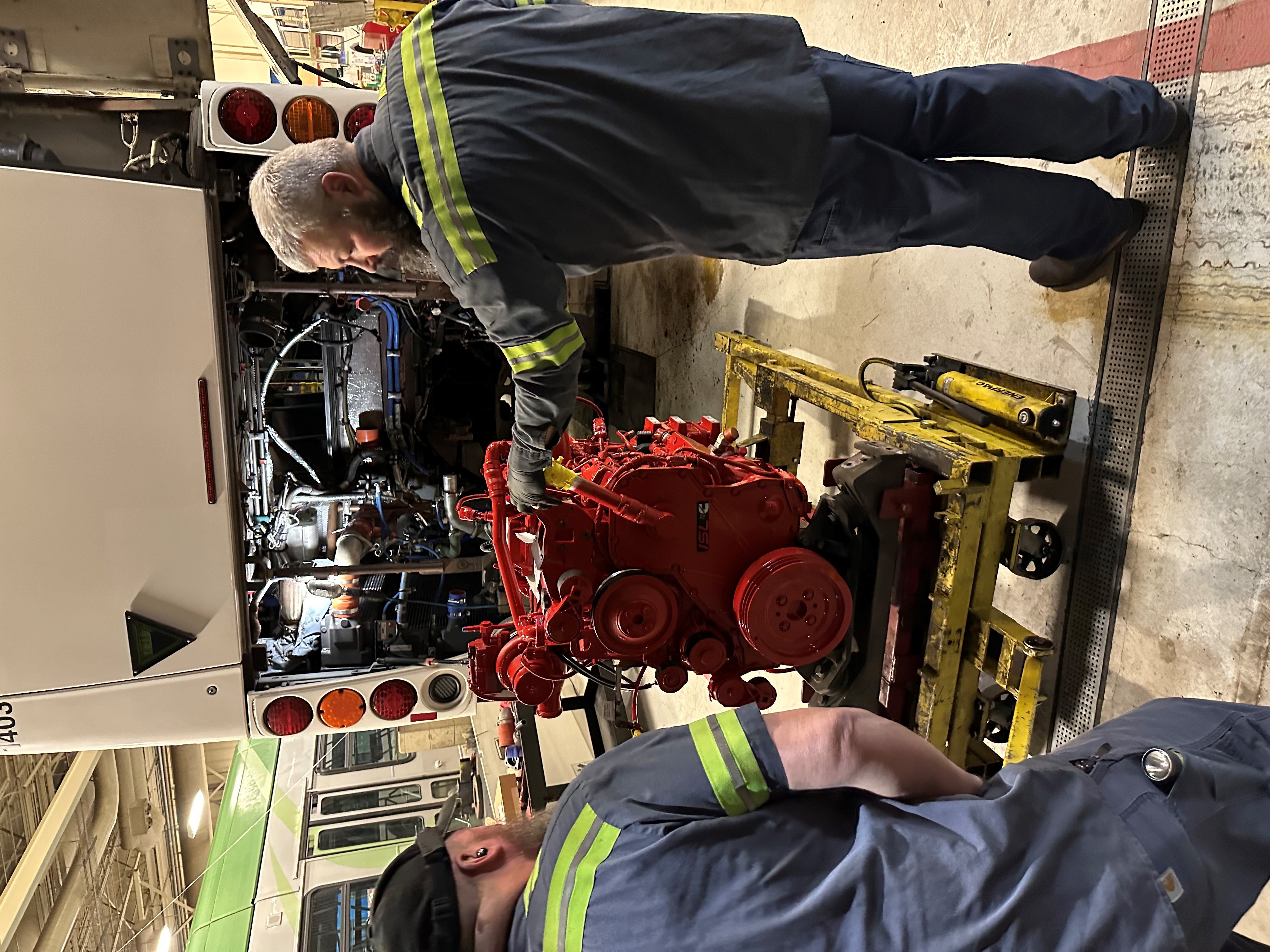 Two mechanics in uniforms stand by a red engine on a lift, preparing to install it into the rear of a bus inside a maintenance garage.