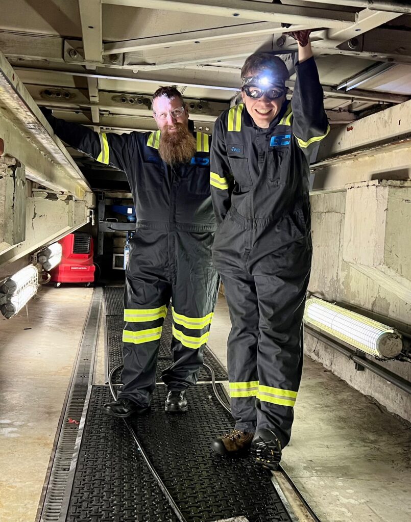Two people wearing dark coveralls with reflective stripes and safety gear stand under industrial equipment in a well-lit mechanical space.