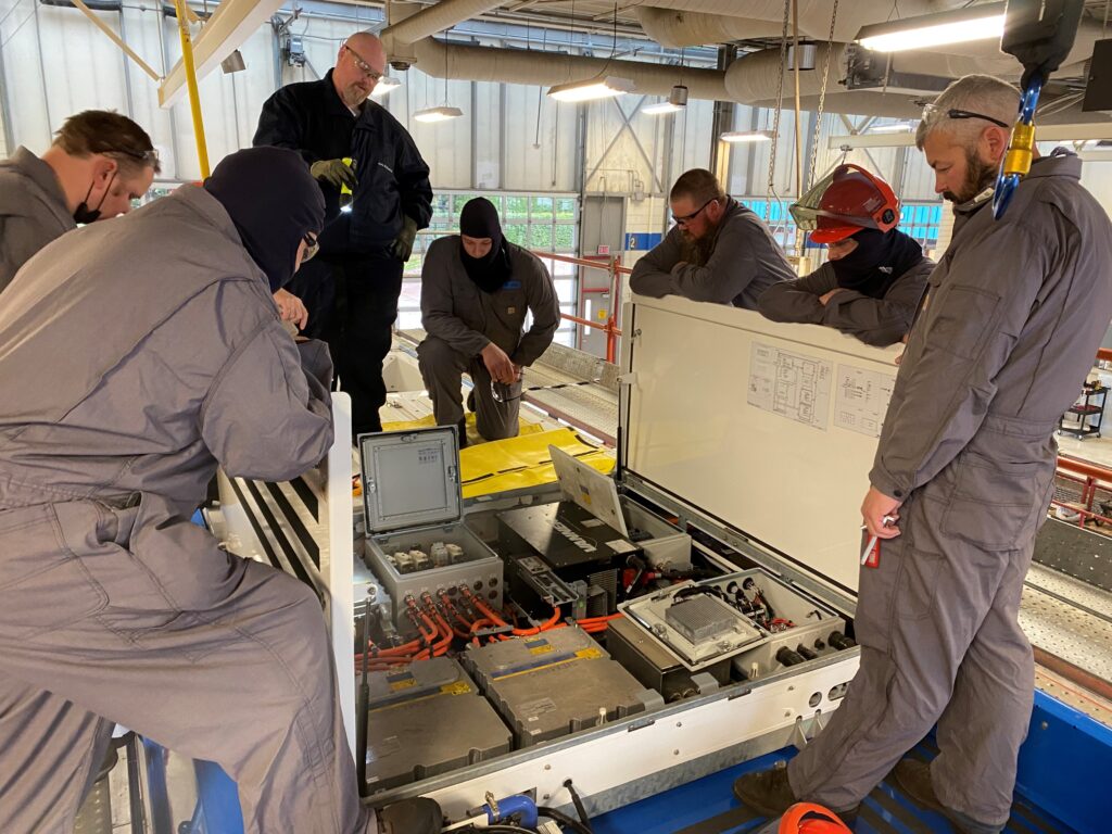 A group of people in gray coveralls and safety gear gather around and inspect an open electrical panel with exposed wiring and components in an industrial setting.