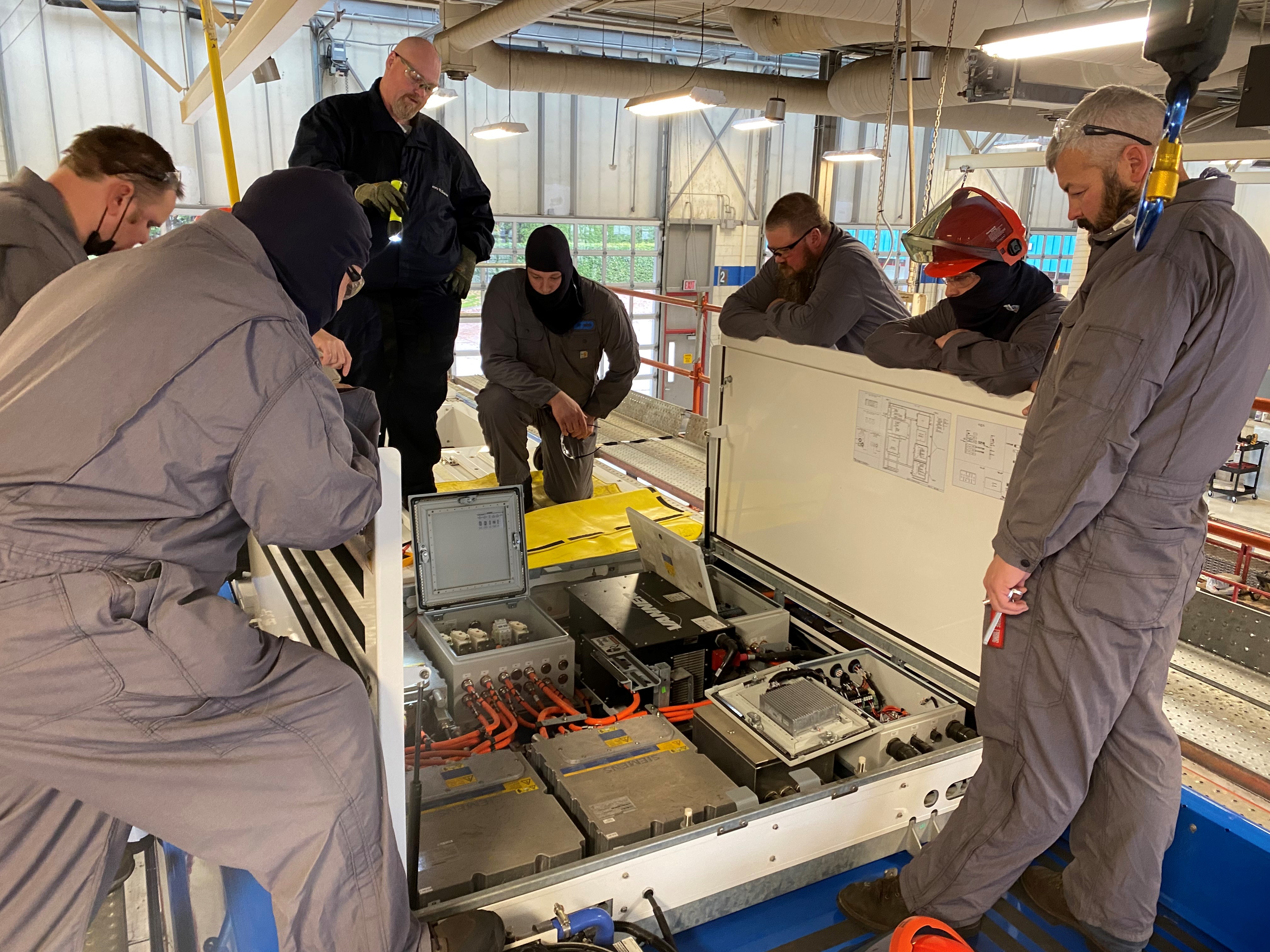 A group of people in gray coveralls and safety gear gather around and inspect an open electrical panel with exposed wiring and components in an industrial setting.