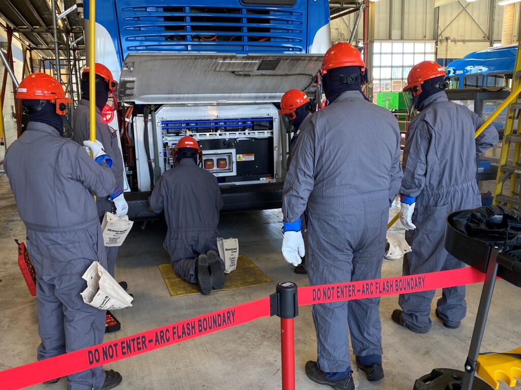 Six workers in protective gear examine electrical equipment at the rear of a bus inside a facility, standing behind a red arc flash boundary warning tape.
