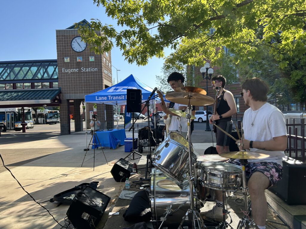 Three musicians perform on drums, guitar, and bass outdoors near Eugene Station, with a blue Lane Transit District tent and clock tower visible in the background.