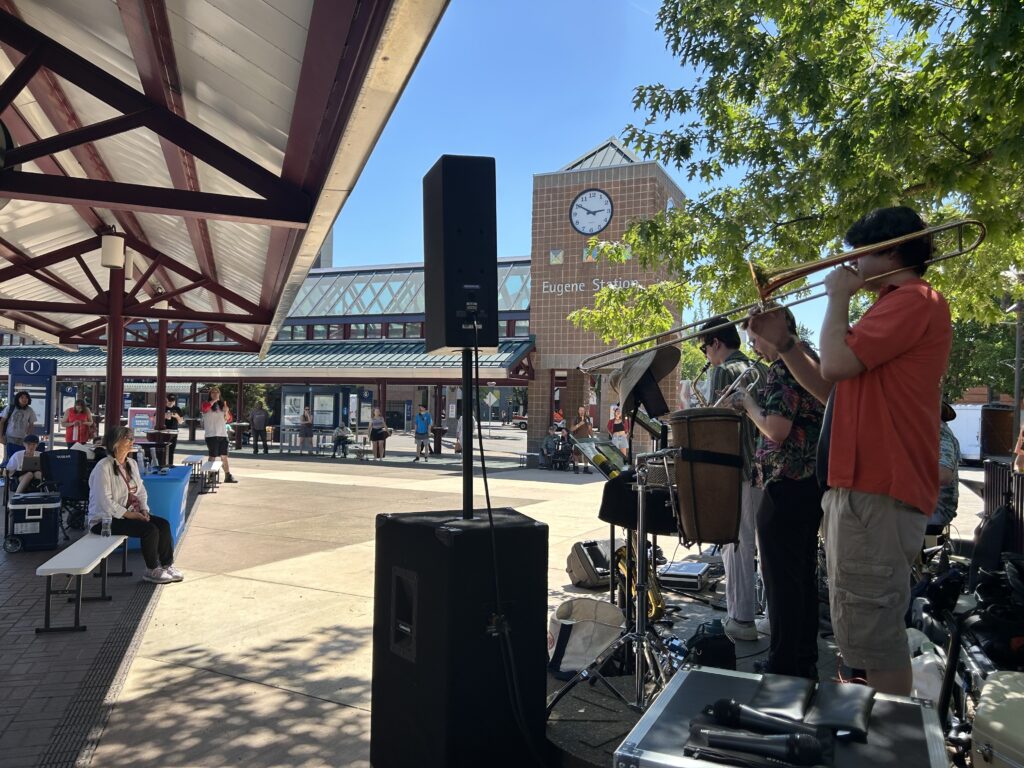 A band performs outdoors near Eugene Station as people sit and watch under a covered area on a sunny day. A large clock and station building are visible in the background.
