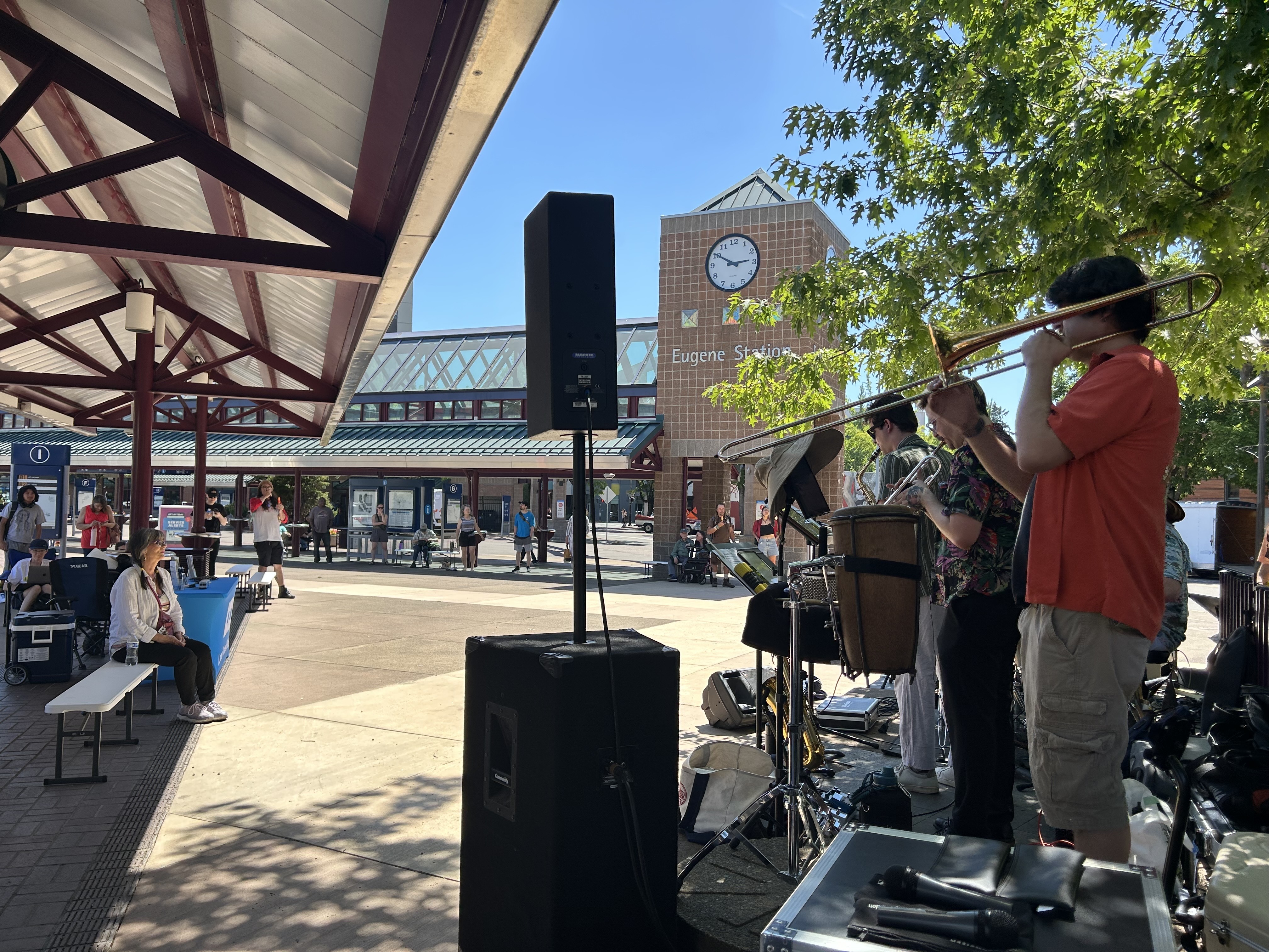A band performs outdoors near Eugene Station as people sit and watch under a covered area on a sunny day. A large clock and station building are visible in the background.