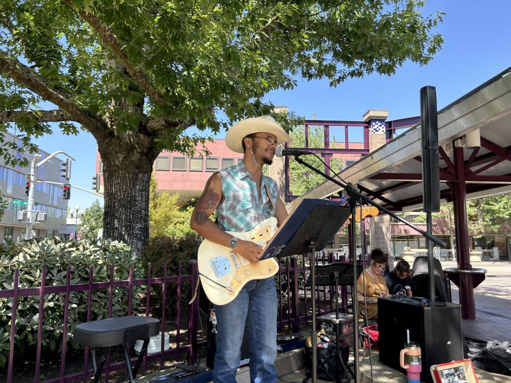A man wearing a cowboy hat and sleeveless shirt plays electric guitar and sings into a microphone at an outdoor performance, with a tree and buildings in the background.