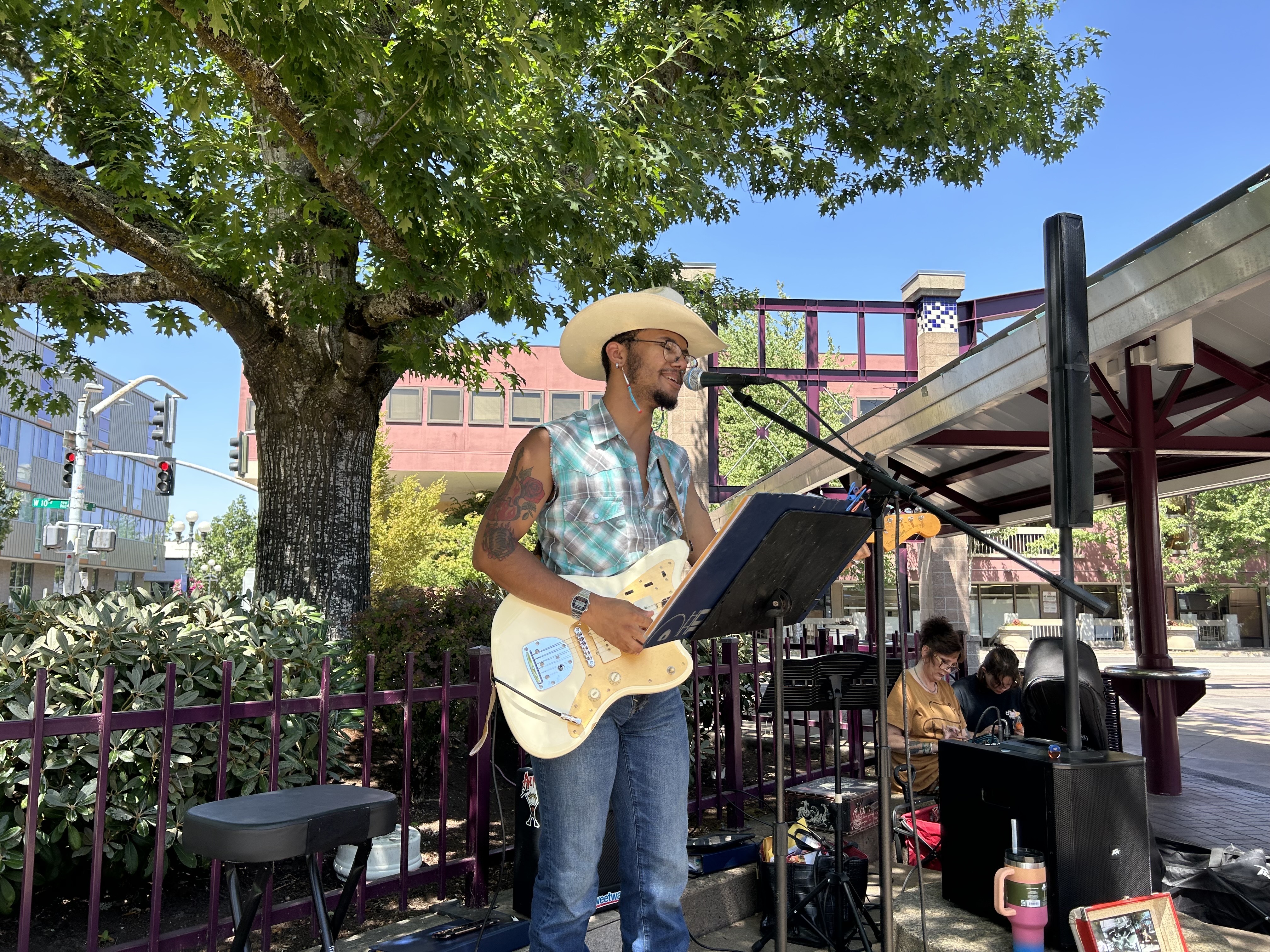 A man wearing a cowboy hat and sleeveless shirt plays electric guitar and sings into a microphone at an outdoor performance, with a tree and buildings in the background.