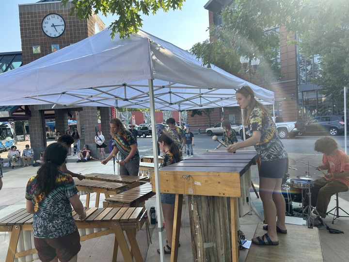 A group of people play marimbas and drums under a canopy at an outdoor event in a downtown area with buildings and a clock in the background.