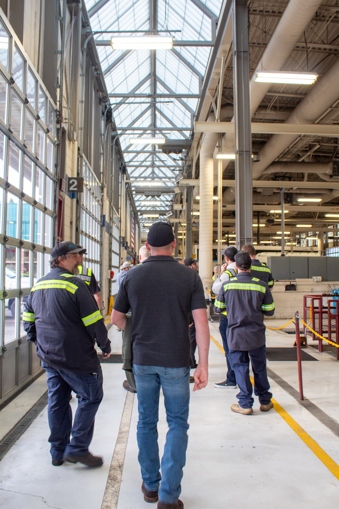 A group of people wearing work uniforms and safety vests walk through an industrial warehouse with high ceilings and large windows.