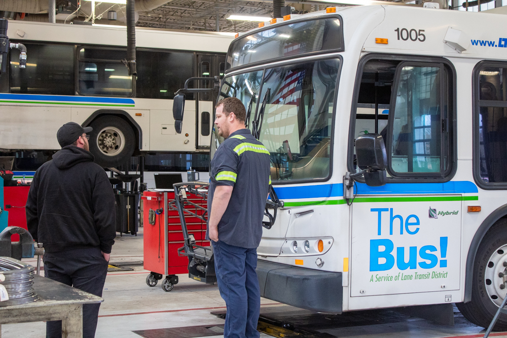 Two men stand next to a city bus in a maintenance garage, with another bus lifted on a platform in the background.