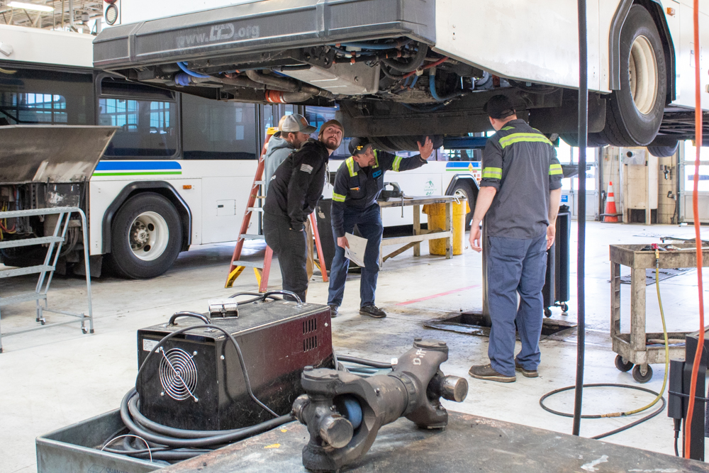 Four mechanics inspect the underside of an elevated bus in a repair garage, with tools and another bus visible in the background.