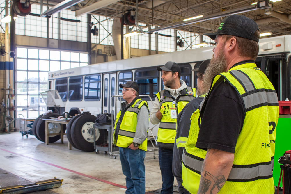 Four people wearing yellow safety vests stand inside a bus maintenance garage, observing a white city bus and equipment in the background.