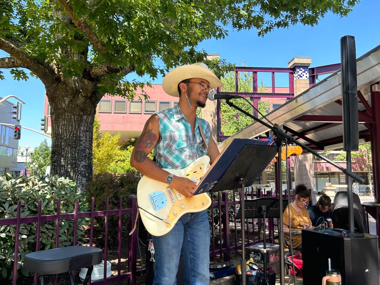 A musician wearing a cowboy hat and sleeveless shirt plays an electric guitar and sings into a microphone at an outdoor venue, with people and music equipment in the background.