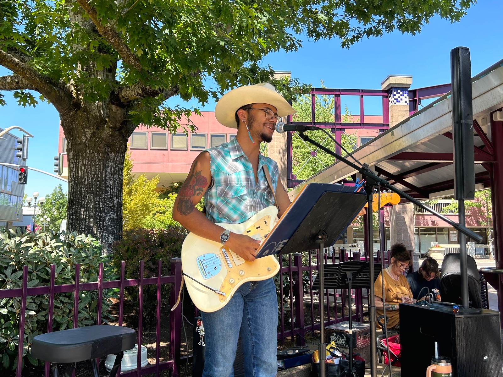 A musician wearing a cowboy hat and sleeveless shirt plays an electric guitar and sings into a microphone at an outdoor venue, with people and music equipment in the background.