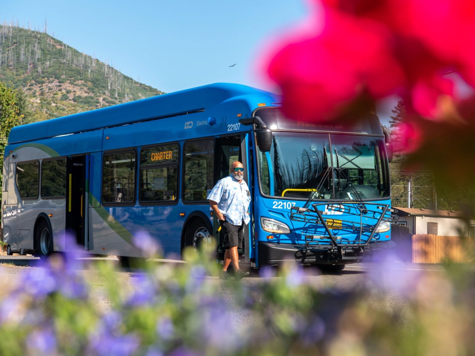 A bus driver stands beside a blue city bus labeled "Charter" on a sunny day, with flowers in the foreground and hills in the background.
