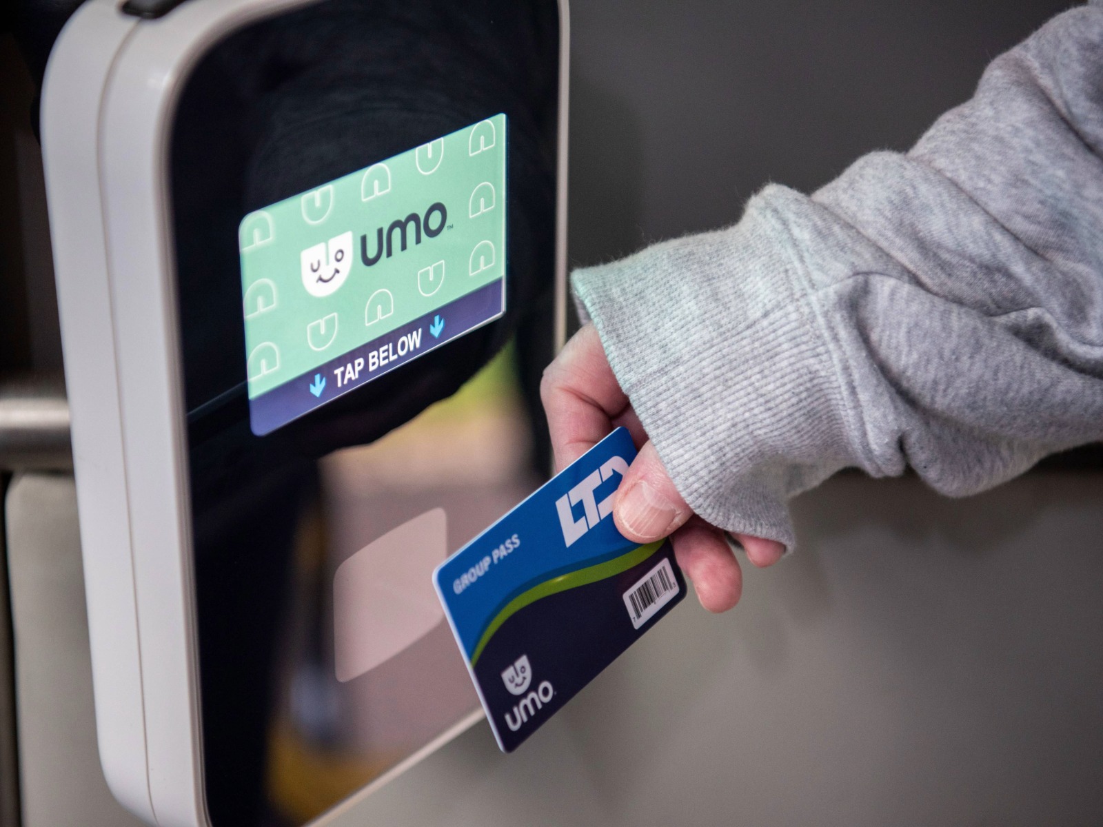 A person holds a Umo group pass card near a contactless payment terminal labeled "TAP BELOW.
