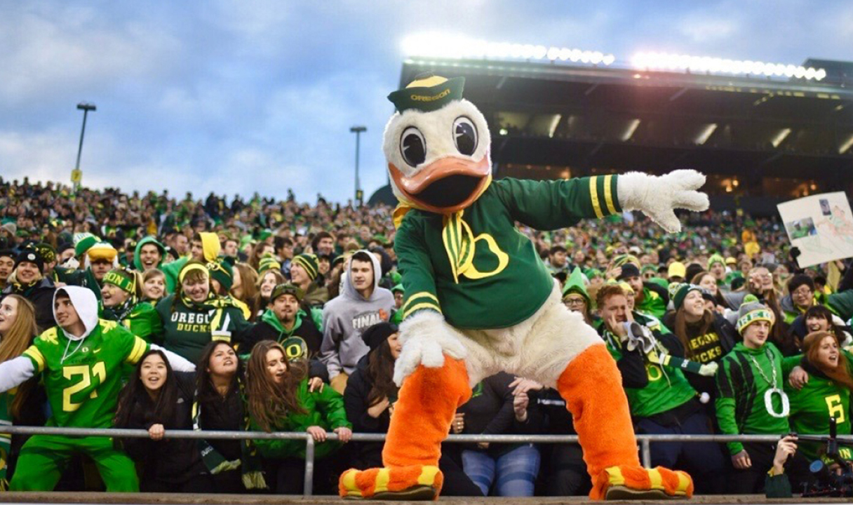 The Oregon Ducks mascot poses in front of a large crowd of fans in a stadium, with people wearing green and yellow team apparel.