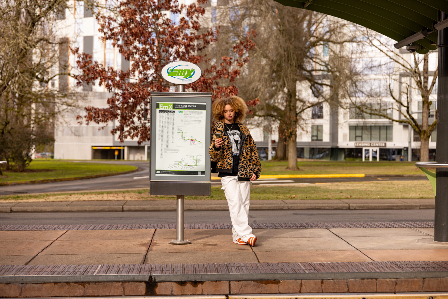 A person stands at a bus stop, looking at their phone next to an EMX transit map, with trees and buildings in the background.