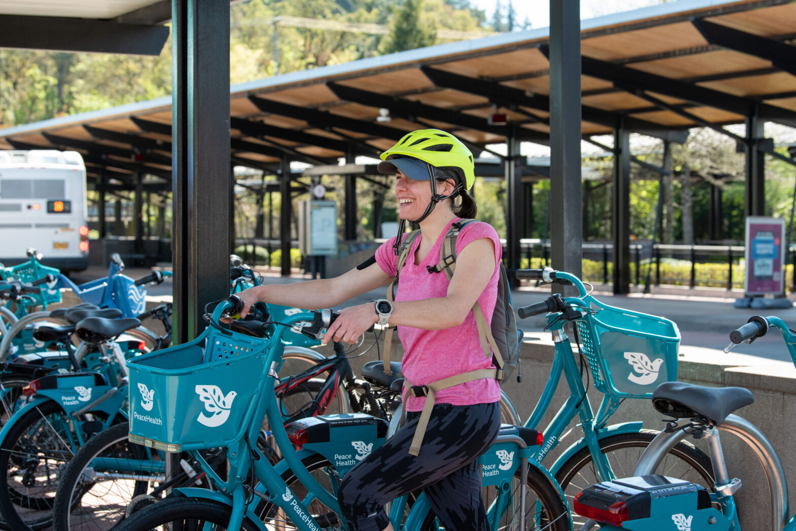 A person wearing a yellow helmet and pink shirt stands next to a row of parked rental bikes at an outdoor bike station.