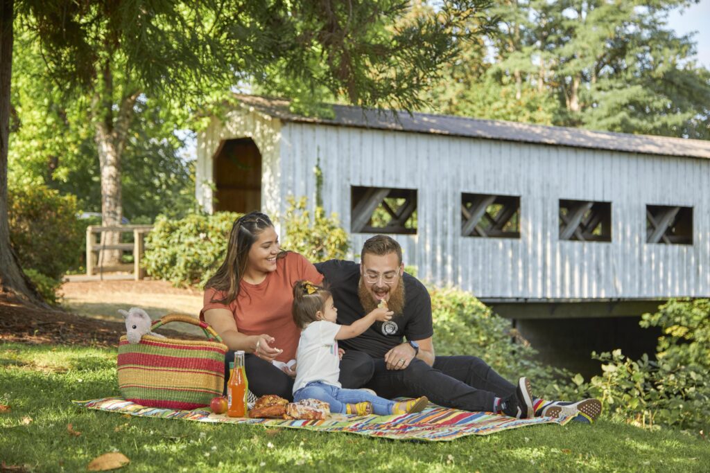 A family of three has a picnic on a blanket near a covered wooden bridge, with food, drinks, and a striped bag on the grass.