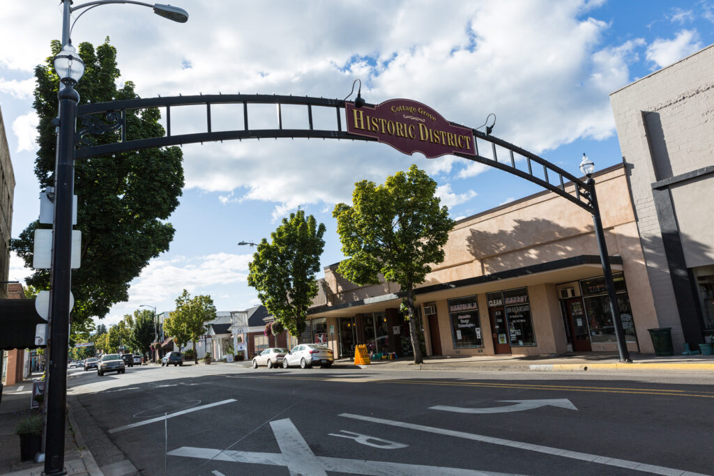 A street view of a small town’s historic district with an arched sign over the road, shops, trees, and partly cloudy sky.
