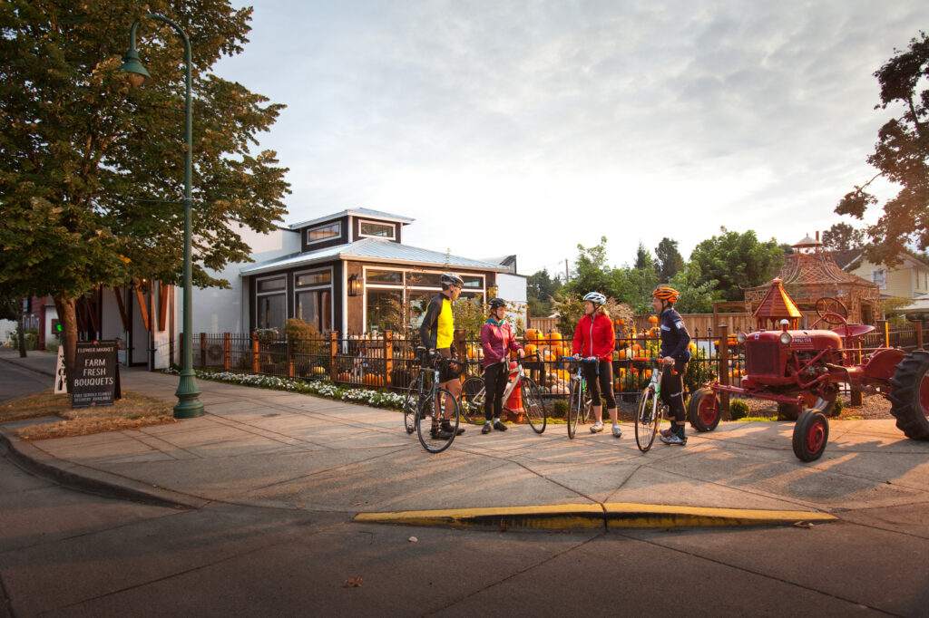 Four cyclists wearing helmets and gear stand with their bikes on a street corner near a café and a red tractor, with trees and a sign that reads “Farm Fresh Roosters” in the background.