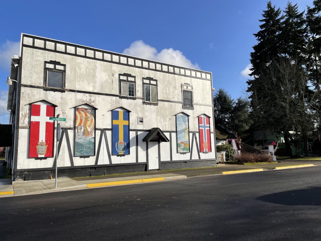 A white two-story building with five large Scandinavian flags and murals displayed on its front facade, bordered by trees and a clear street.