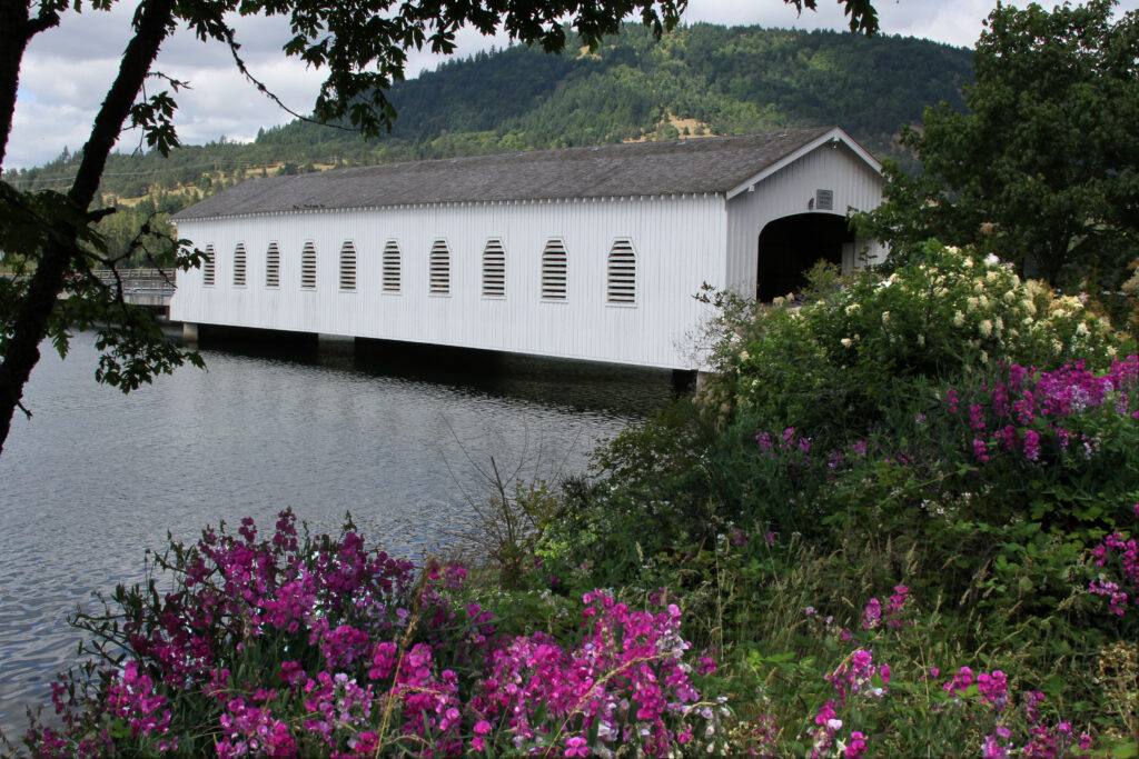 White covered bridge spans a river, surrounded by lush greenery and blooming purple flowers, with hills in the background under a partly cloudy sky.