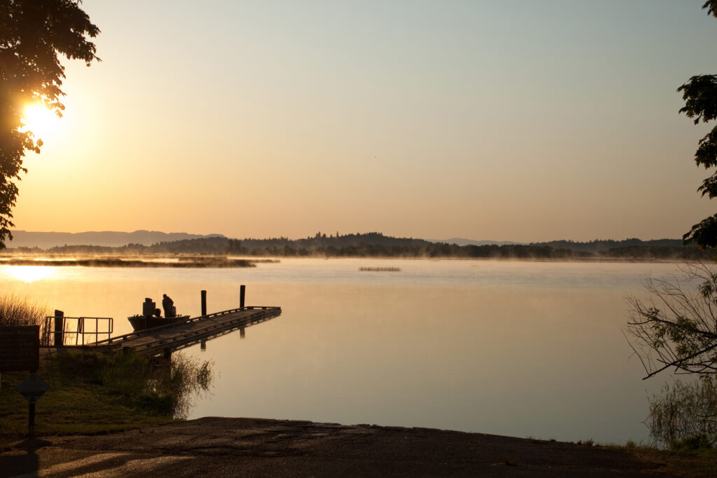 A dock extends into a calm lake at sunrise, with two people sitting at the end. Mist rises from the water and trees frame the scene.