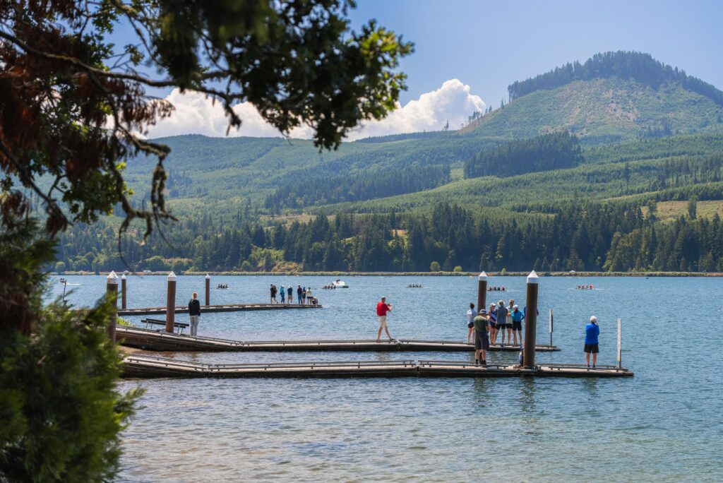 People stand and walk on docks over a lake, with forested mountains in the background under a partly cloudy sky.