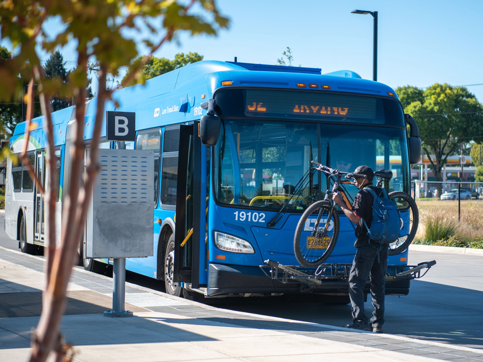 A person loads a bicycle onto the front rack of a blue city bus at a transit station on a sunny day.