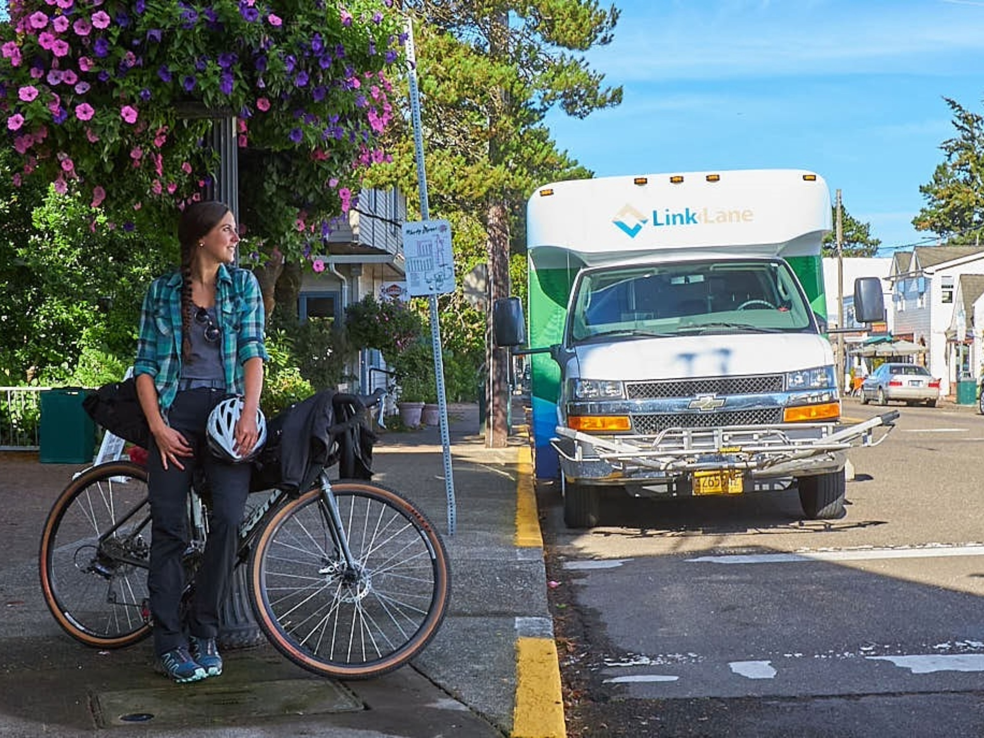 A person with a helmet sits on a bicycle near a sidewalk, looking away. A Link Lane shuttle bus is parked on the street in the background.